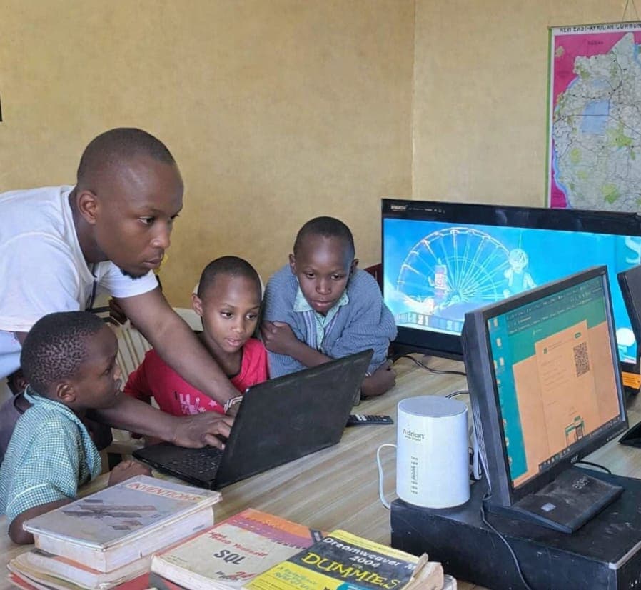 Students gathered around a laptop as a facilitator demonstrates digital skills.